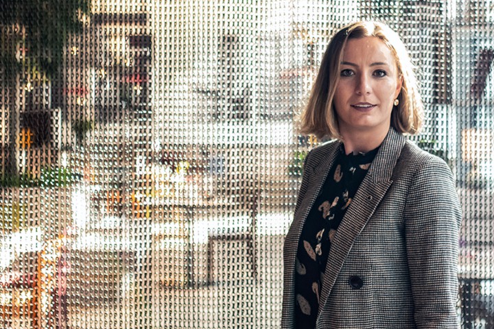 In a light and modern hotel lounge area. A young woman in a business outfit is looking directly into the camera.