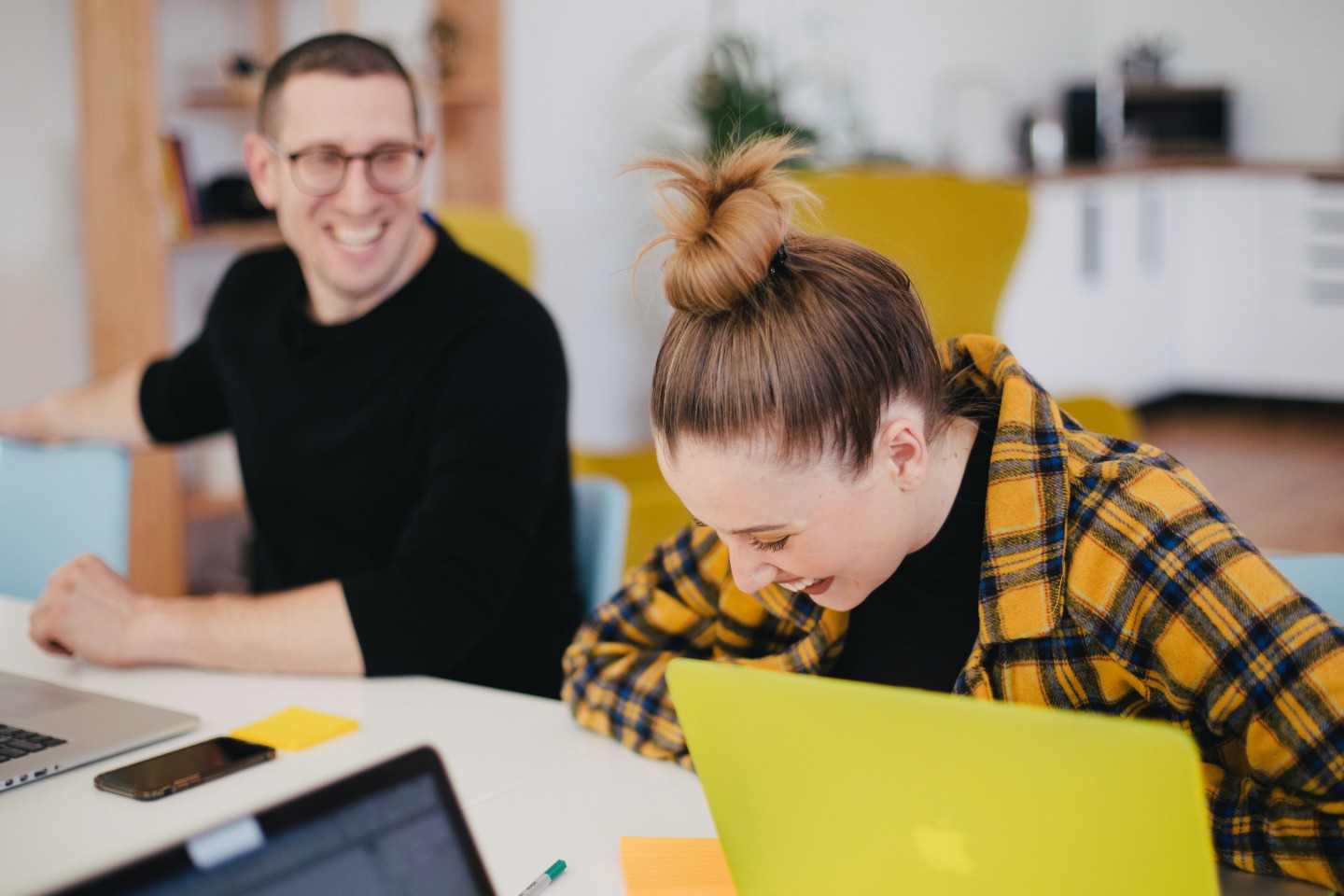 Two young people are sitting at a conference table in front of their laptops. Both are laughing. They wear casual clothes.