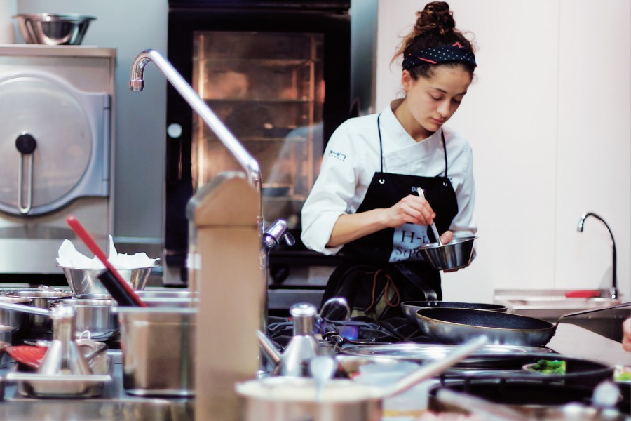 In a large hotel kitchen. A young chef is stirring food in a small pot.