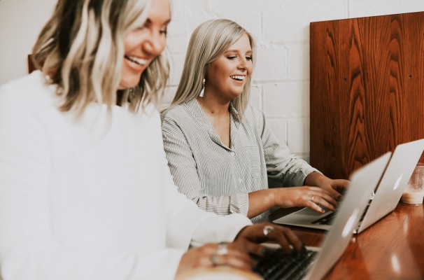 Two laughing employees are working side by side on their modern laptops.